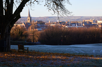 Tapton park winter spire This landscape photograph captures Tapton Park in Chesterfield, United Kingdom, on a frosty winter morning. The scene is dominated by nature, with trees displaying sparse winter foliage and a layer of frost covering the park grounds. A wooden bench sits beneath a large tree in the foreground, with scattered brown leaves on the ground. In the distance, the prominent spire of the Church of St Mary and All Saints rises above the town's buildings, serving as a distinctive landmark and adding architectural interest to the image. The park provides a tranquil setting overlooking Chesterfield, and the winter sunlight highlights the texture of the frost and the natural beauty of the surroundings. This view effectively showcases both the historic church and the peaceful landscape of Tapton Park.